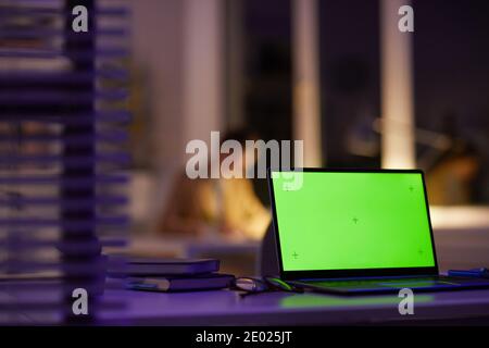 Modern office workplace late in evening with green screen laptop on foreground and unrecognizable woman working on background, copy space Stock Photo