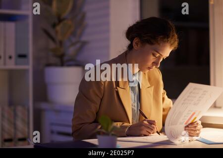 Serious young woman concentrated on paperwork routine sitting at desk checking finance data late in evening Stock Photo