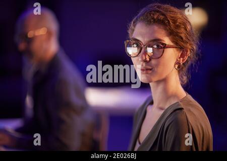 Medium close-up portrait shot of beautiful young woman wearing eyeglasses working in office in evening looking at camera Stock Photo