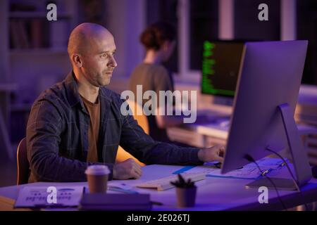 Handsome Caucasian man sitting at desk in front of computer monitor working on computer program in evening Stock Photo