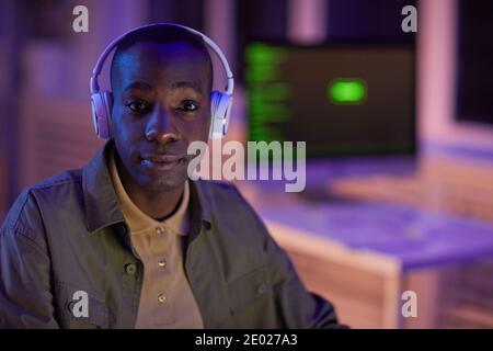 Medium close-up portrait of handsome young adult Black man wearing casual outfit and headphones spending evening in modern office looking at camera Stock Photo