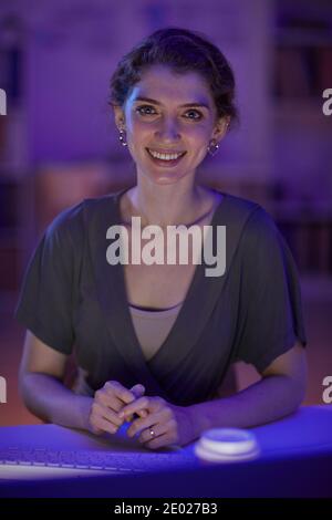 Vertical medium portrait of attractive young Caucasian woman sitting at desk in office room late in evening smiling at camera Stock Photo