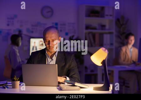 Group of three modern office workers sitting at desks finishing workday late in evening, copy space Stock Photo