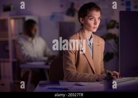 Portrait of serious young woman wearing elegant outfit sitting in front of computer focused on work in evening Stock Photo