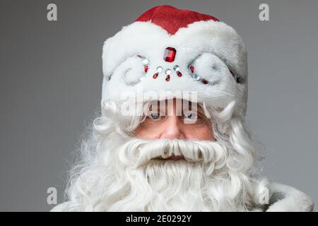 Headshot of Santa Claus in red hat, gray-haired beared tired man on gray studio background Stock Photo