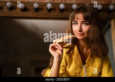 Portrait of a young beautiful woman in yellow bites a slice of pizza close-up Stock Photo
