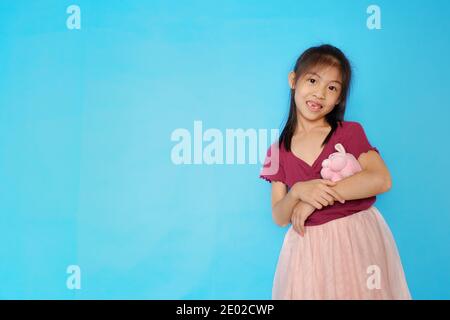 A closeup picture of a cute Asian girl, looking straight at the camera, smiling, cradling her pink pig doll with plain light blue background. Stock Photo