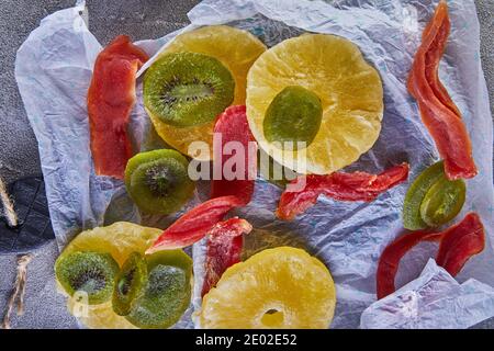 Dried fruits: Yellow candied pineapple rings, red papaya and green on white wrapping paper on gray concrete Stock Photo