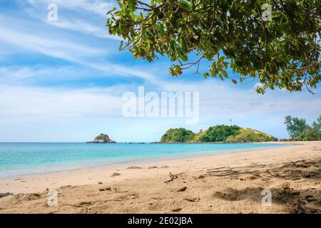 View of a deserted tropical beach. Yellow sand, turquoise sea and blue sky with clouds. On the horizon are green hills with a white lighthouse at the Stock Photo