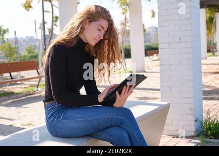 Redhead caucasian woman reading or working in a e-book or tablet in a park. Digital nomad entrepreneur concept. Stock Photo