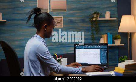 African freelancer analysing digital blue prints checking project of company looking on laptop sitting at desk in living room office late at night overtime. Busy black employee using modern technology Stock Photo