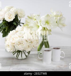 White roses, lily, in round vases with two cups of coffee on the table for a special occasion as a kitchen decoration. Copy space Stock Photo
