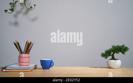 Blue ceramic mug, pencil holder, colored pencils, notebooks and a pot of small pine tree on table. Home work desktop design composition. Stock Photo