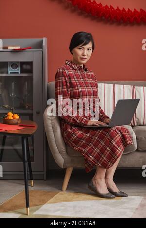 Beautiful mature woman wearing stylish red dress sitting on sofa in living room surfing Internet using laptop looking at camera, full shot Stock Photo