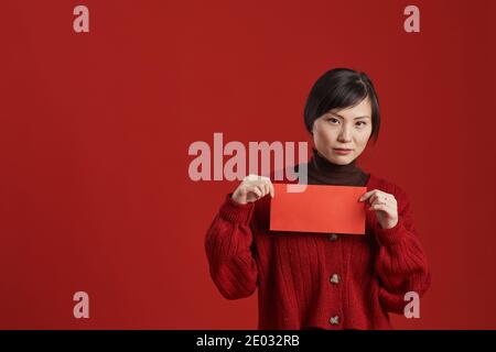 Medium studio portrait shot of attractive Asian woman wearing stylish outfit holding red envelope standing against red background, copy space Stock Photo