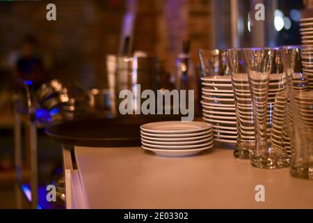 Stacks of clean dishes on the table of the bollard. White ceramic plates and saucers, glass transparent glasses. Stock Photo