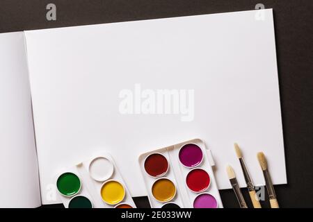Watercolor paints, brushes and an open sketchbook on a dark background with copy space, top view. Stock Photo