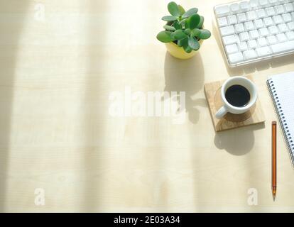 Top view of wooden desk in office workplace with office supplies in sunlight. Keyboard, notepad, pen, potted plant, and coffee. Copy space Stock Photo