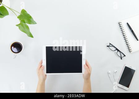 Human's hands holding digital tablet on modern office desk. Top view of workspace with coffee, eyeglasses, smartphone, earphone, notepad, pen and gree Stock Photo