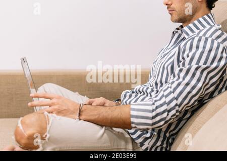 Detail of the body of a man working sitting on the couch at home with a laptop between his legs Stock Photo