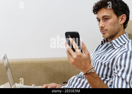 Portrait of a man working with a laptop while using the mobile on the couch at home with a laptop in his legs Stock Photo