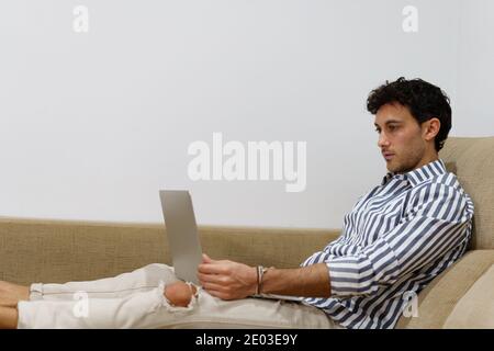 Portrait of a young man working with a laptop while lying on the couch at home with a laptop in his legs Stock Photo