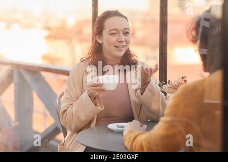 Young woman drinking coffee and talking to her friend during their meeting in cafe Stock Photo