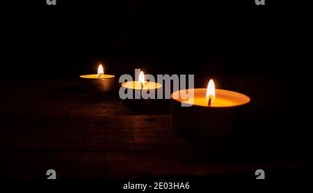 Three lighted tea wax candles on a wooden table on a black background. Stock Photo
