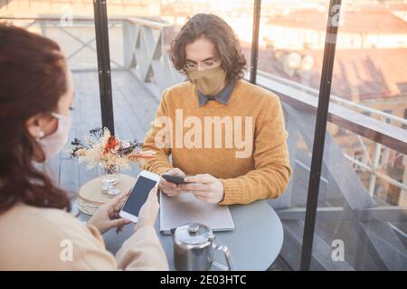 Young couple in masks using their mobile phones while have meeting in coffee shop during pandemic Stock Photo
