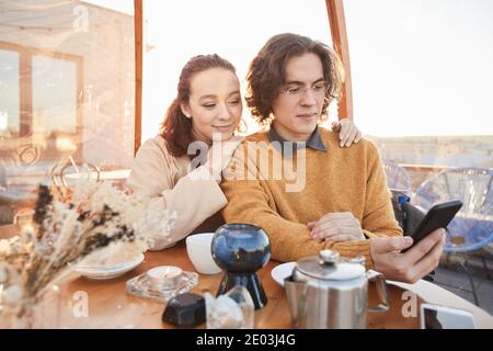Young couple watching video online on mobile phone while drinking tea in coffee shop Stock Photo