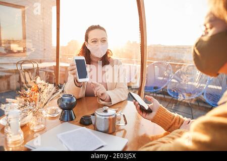 Two business colleagues in masks discussing online work using their mobile phones during business meeting in cafe Stock Photo
