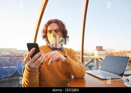 Young businessman in mask using his mobile phone while sitting at the table with laptop and working in coffee shop Stock Photo