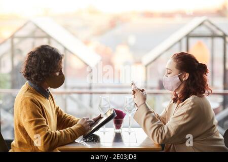 Two business partners in masks working online using mobile phone and tablet pc in cafe Stock Photo