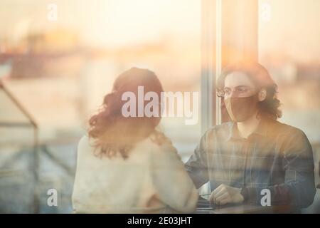 Two business colleagues in masks sitting at the table behind the window of cafe and talking to each other Stock Photo