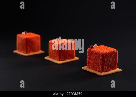 Closeup view of matte orange dessert decorated with chocolate balls isolated on black studio background. Three small square matte cakes in row on cookies in restaurant. Concept of confectionary. Stock Photo