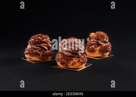 Three delicious fresh crunchy profiteroles with sweet brown chocolate creamy filling. Closeup of homemade tasty baked eclairs isolated on black background. Concept of desserts, restaurant food. Stock Photo