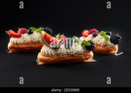 Side view of dessrt filled with cream, white topping and slices of figs, blackberries, raspberries and blueberries. Fresh three baked homemade eclairs in row isolated on black background. Stock Photo