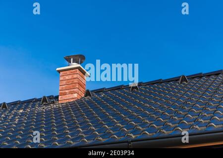 New chimney pipe in red bricks on roof in black shingles. On blue sky. Construction of a family house. Detail of the roof. Stock Photo