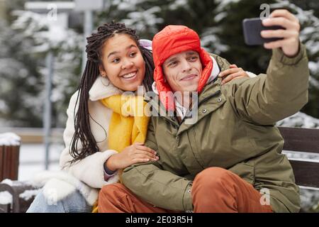 Young man making selfie portrait on his mobile phone with woman while they sitting on bench in winter park Stock Photo
