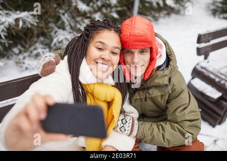 Young happy woman posing at camera of her mobile phone together with man while they sitting on bench in winter Stock Photo