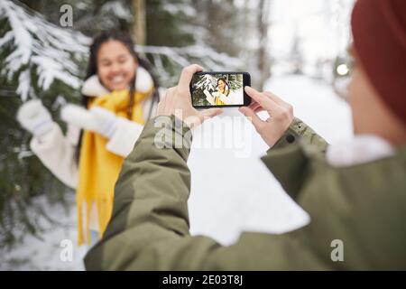 Close-up of man photographing woman on mobile phone while she posing against the snowy trees outdoors Stock Photo