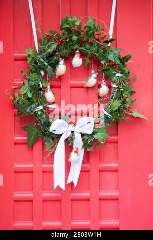 Home-made Christmas wreath on a red door. The wreath uses holly, ivy and rosemary leaves, white ribbon and bird-shaped baubles. Anna Watson/Alamy Stock Photo