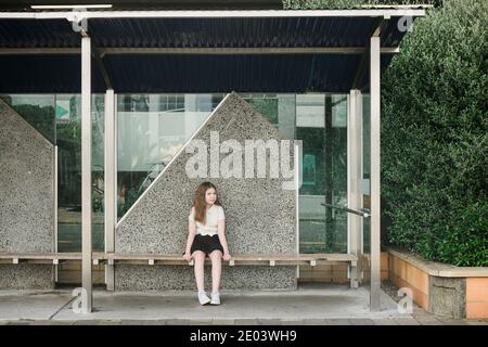 Young girl sitting alone on a wooden seat at an empty bus stop Stock Photo