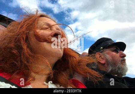 Elderly couple sitting in a convertib. The man wears a cap and is the driver. The wind is gripping the red hair of the lady.  Blue sky with clouds Stock Photo