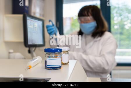 Female clinical laboratory scientist in protective face mask and glasses holding pipette. Close-up of glass vial and syringe on lab analyzer. Covid-19. Stock Photo