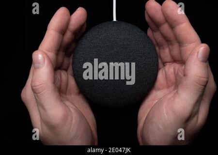London, United Kingdom - 19 December 2020: Hands around a Google Nest Home Mini smart speaker with built-in Google Assistant on a black background. Stock Photo
