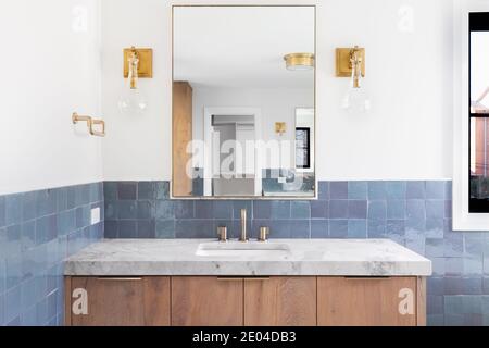 A luxury home's renovated bathroom with blue tiles, a light wood vanity cabinet, gold lights mounted on the wall, and a grey stone counter top. Stock Photo