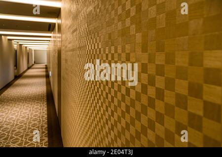 A shimmering checkerboard wood marquetry lines the wall of a long corridor at The Okura Tokyo, Minato ward, Japan. The carpet features a geometric diamond pattern in beige and brown. Stock Photo