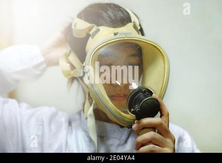 A Asian woman pulling on a full face protective oxygen mask, white coat and rubber gloves, getting ready to work at a biochemical lab. Stock Photo