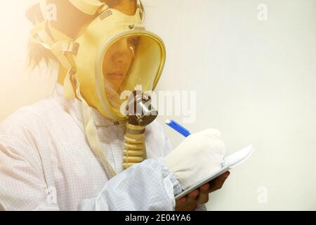 A woman with a full face protective oxygen mask, white coat and rubber gloves, getting ready to work at a biochemical lab, taking notes with her pen a Stock Photo